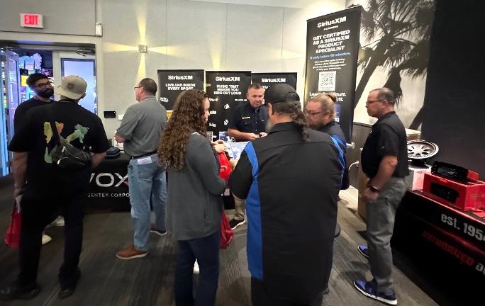 A group of people gathered around a booth at an event, engaging with staff and discussing products. The booth features banners for SiriusXM and a display of audio equipment.