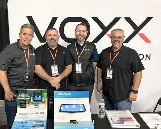 Four men posing together at a booth displaying VOXX products, including electronics and accessories.