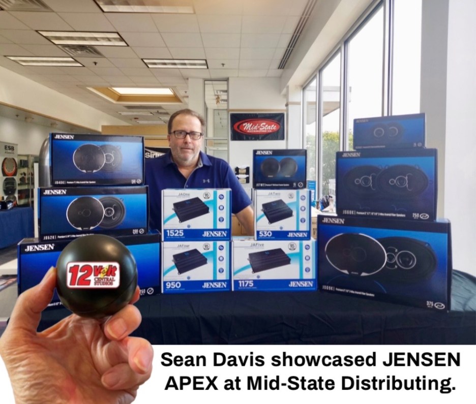 A person stands behind a table displaying various JENSEN audio products at a distribution center, holding a black ball with '12 Volt' written on it.