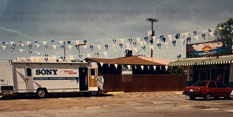 A Sony Road Show trailer parked in a lot, decorated with blue and white triangular flags, alongside a red car and a shop with a sun mural.