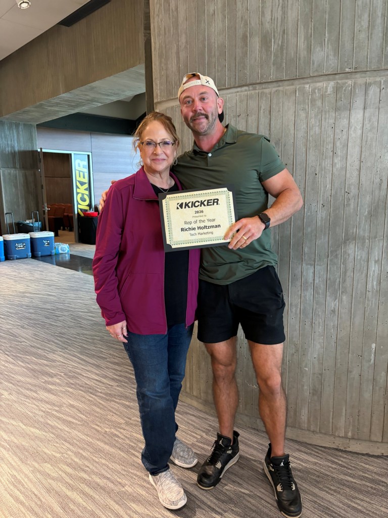 A man and a woman posing together inside a modern building, holding an award certificate. The man is wearing a green polo shirt and black shorts, while the woman is wearing a maroon jacket and blue jeans.
